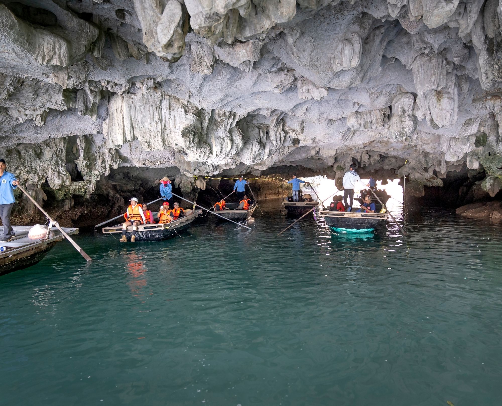 Tourists taking a bamboo boat rowing trip through Luon Cave in Halong Bay, featuring low limestone ceilings and emerald water